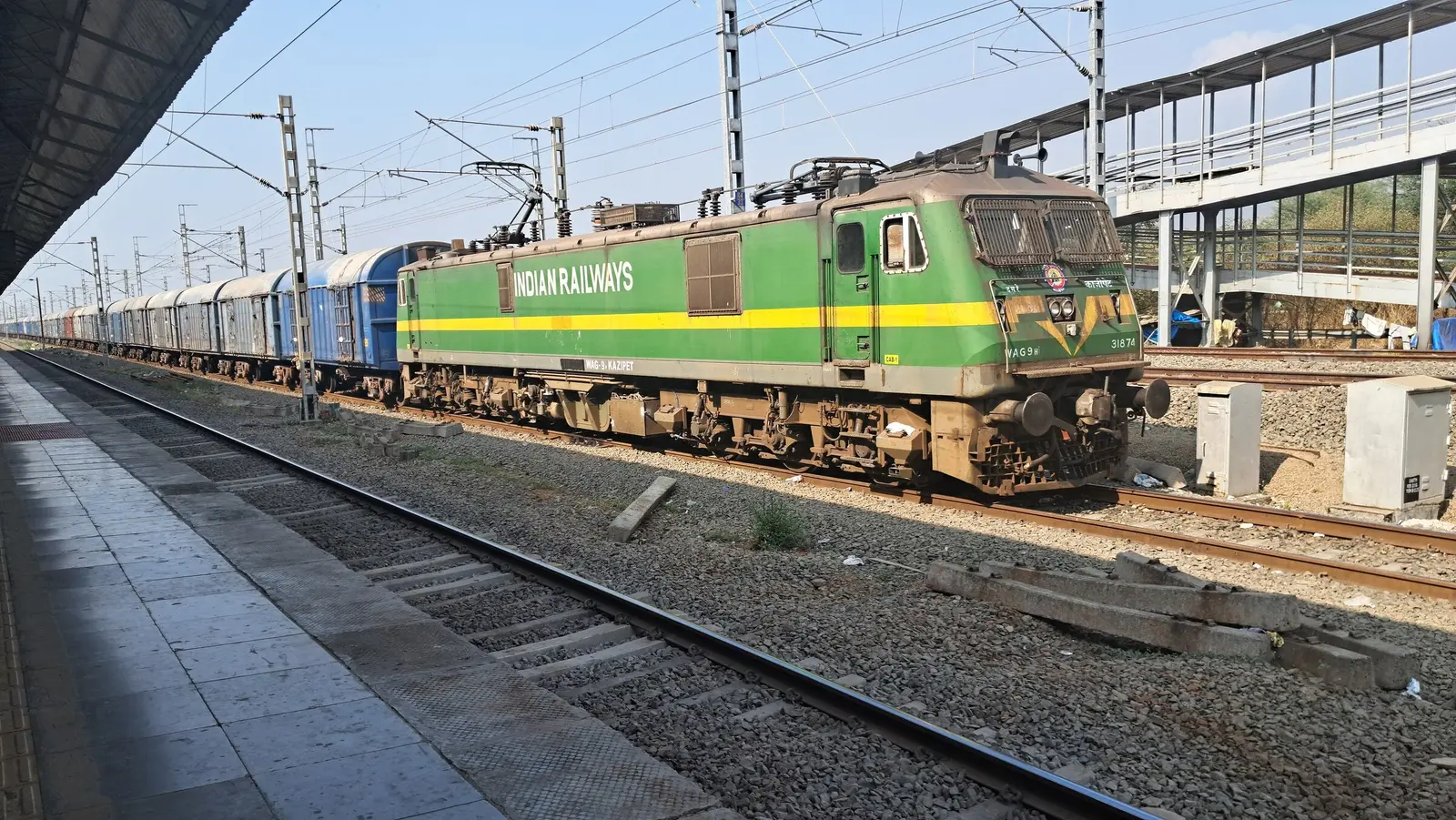 A green WAG-9 electric locomotive hauling a goods train past a station platform